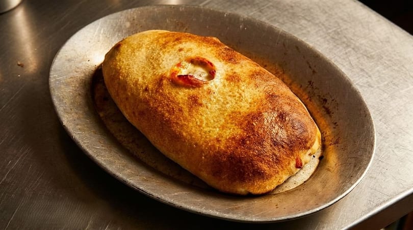 Golden-brown calzone with a small opening revealing cheese and tomato, served on a metal plate.
